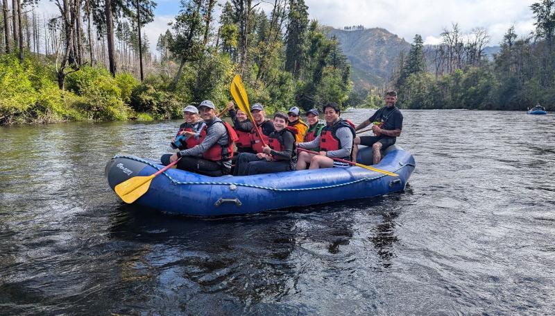 Several people on a blue raft floating on a calm river with forest behind them.