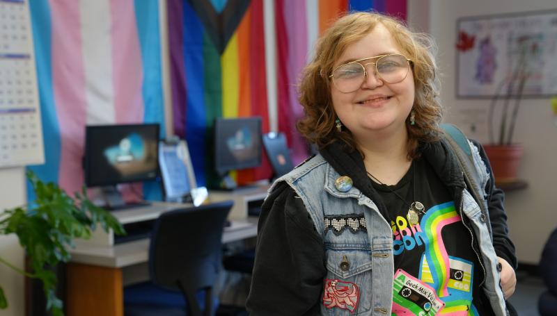 A student with a warm smile, big glasses, and a jean jacket with pins in the Gender Equity Center with pride flags in background