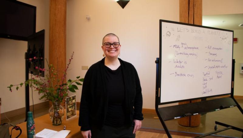 Sci Fi author Becky Chambers smiling and standing next to a whiteboard reading "Let's Build a Planet"