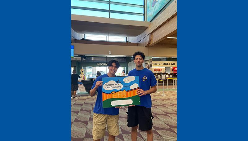 Two smiling LCC employees hold up a welcome sign at the Eugene airport for international students
