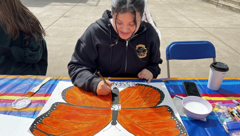 a young Latina woman in a MEChA sweatshirt paints protest sign with a monarch butterfly and words "migration is essential"