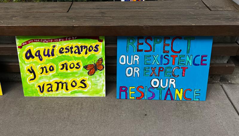 colorful protest signs that read "aqui estamos y no nos vamos" and "respect our existence or expect our resistance"