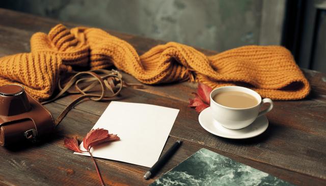 a cup of coffee, some fall leaves, a scarf, a notepad, pen, and vintage camera laid out on a wooden table