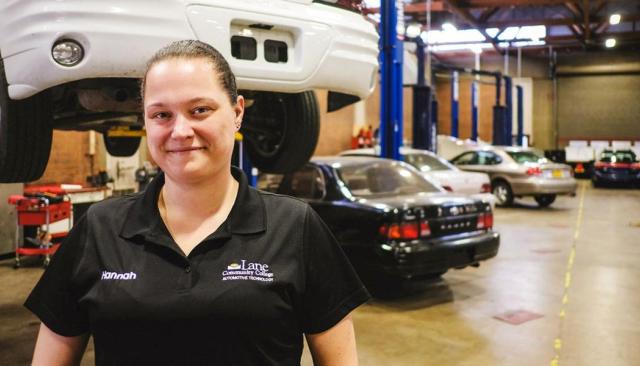 a smiling automotive tech student stands in front of a car on a lift