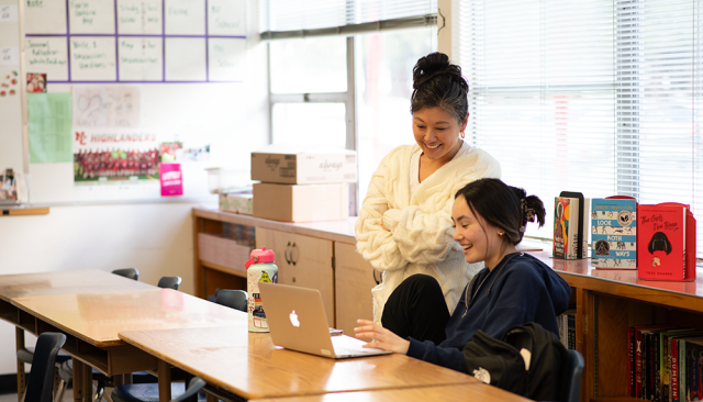 A teacher works with a smiling student on a laptop with displays of children's books in background