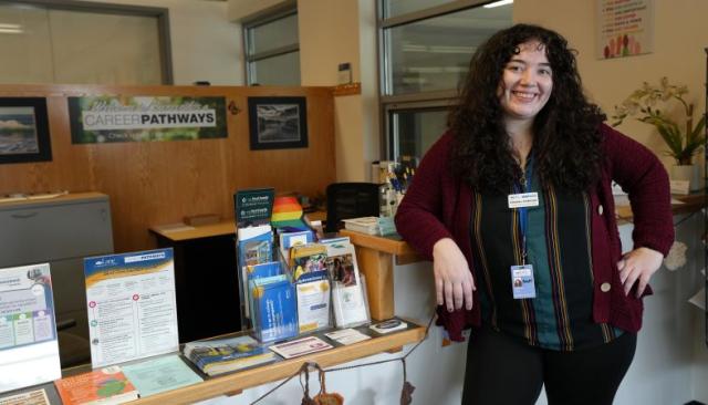 Career Pathways admin Chelsey Andersen smiles warmly next to a desk with a rainbow pride flag and career resource pamphlets