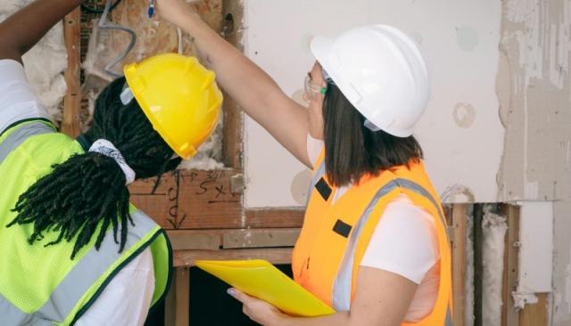 Two electricians in hard hats and vests inspect wiring at a job site
