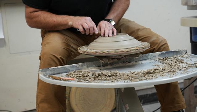 an artist throwing clay on a wheel, carving the bottom of a bowl
