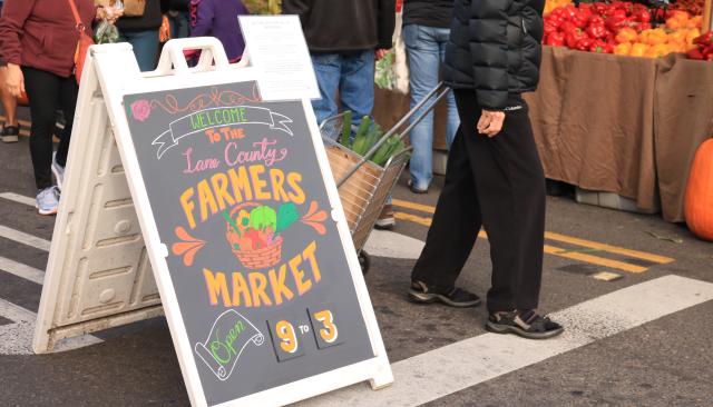 a colorful sign reading "welcome to the Lane County's Farmers Market" with people and produce in the background