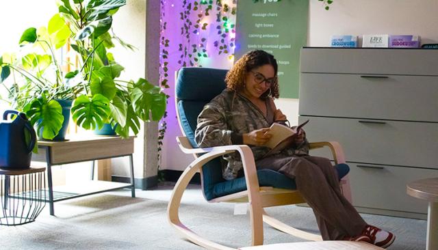 A student, a Black woman in a camo jacket, sits in a comfy chair by a bright window, smiling and reading