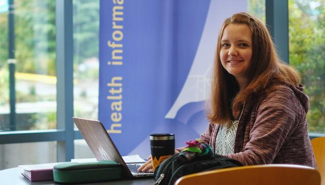 Pre-professional health professions student with a laptop smiles in a sunny lobby