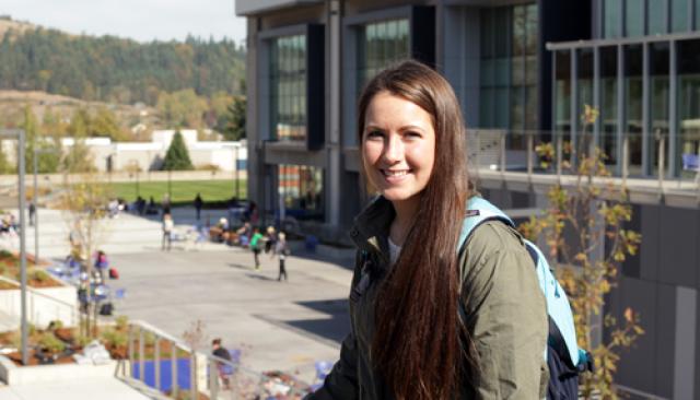 A young woman with a backpack smiling in front of Center Building on main campus