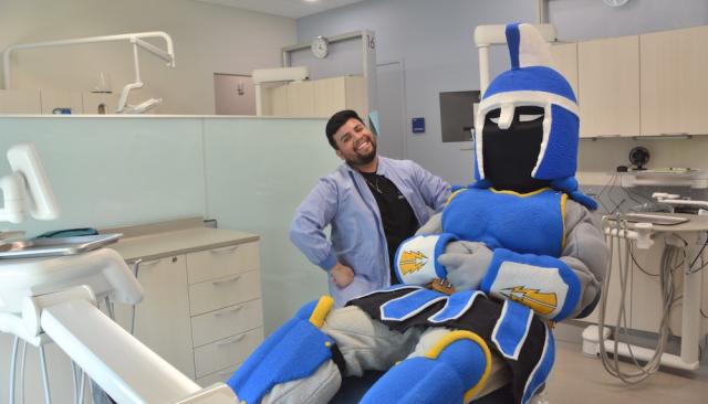 Ty the Titan sitting in an exam chair in the new dental clinic next to a smiling dental hygiene student, a young man of color