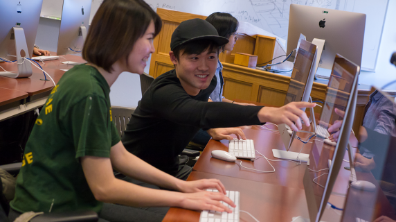Two international students, a young Asian man and woman, smile and work together in a computer lab