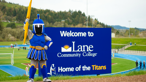 Ty the Titan mascot holds up a lightning bolt by a big LCC sign next to the track