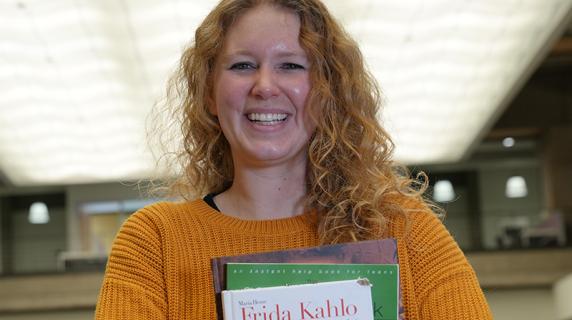 a smiling young woman in a big yellow sweater holding textbooks in the library