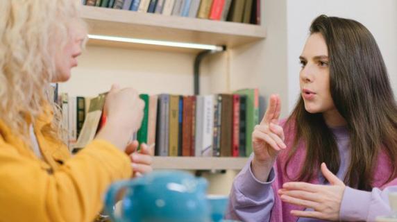 two young women, students in a library, talk together with American Sign Language