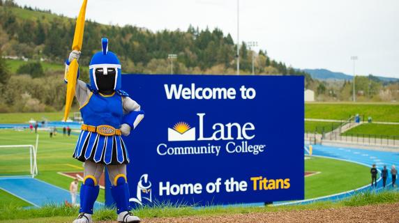 Ty the Titan mascot holds up a lightning bolt by a big LCC sign next to the track