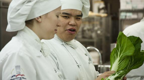 two culinary students talking in a kitchen one with lettuce in their hand 