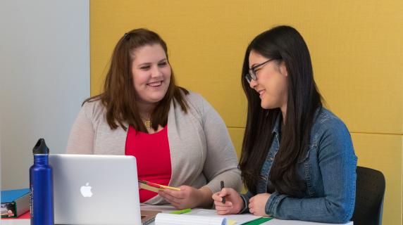 Two students working together at a table with a laptop and papers scattered about