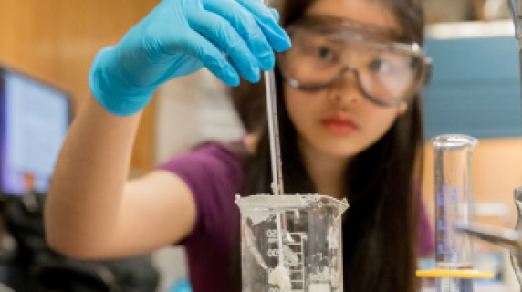 A female student of Asian descent with protectie gear uses a beaker in a chemistry class