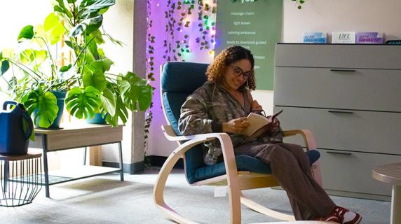 A student, a Black woman in a camo jacket, sits in a comfy chair by a bright window, smiling and reading