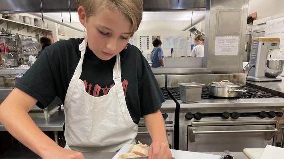 a middle schooler in an ACDC shirt chops chicken at a culinary summer camp