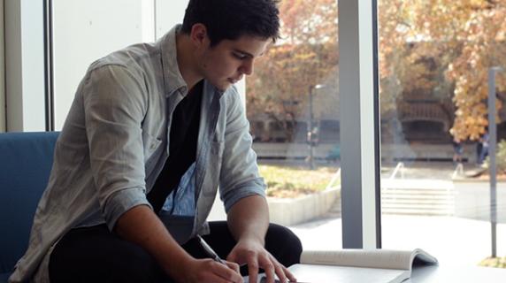 a young man sits by a window in the center building doing homework, with fall foliage outside
