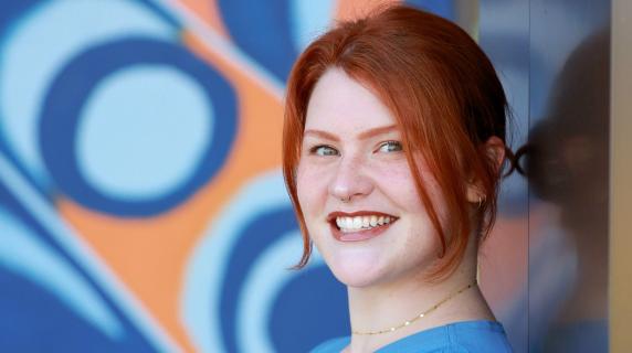 Abby, red haired person wearing blue scrubs leaning against a mural of abstract artwork.