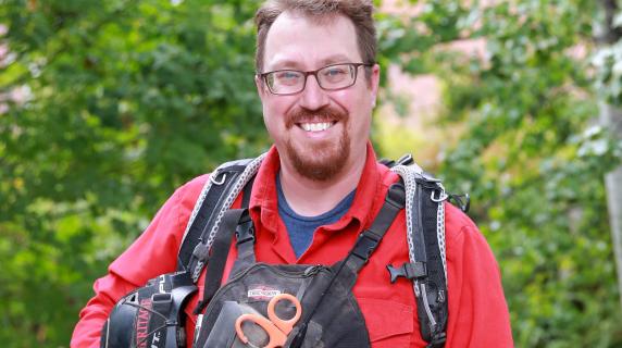 David, a smiling student wearing a red shirt, hiking backpack, and utility vest stands on a wooden path surrounded by green trees, holding a helmet under one arm.