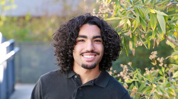 Jamari, a curly haired student leaning on an arm rail with his other had in his pocket smiling at the camera
