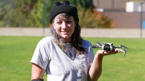 Kayla,A smiling person stands in a grassy field holding a small drone in one hand, wearing a black beanie, white shirt, and dark pants.