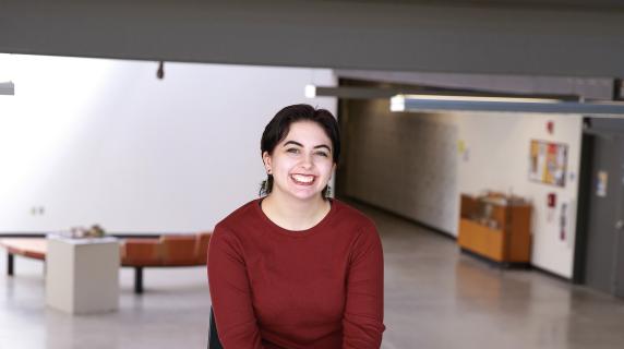 Lily, a student sitting in a chair in  building 10 where a red shirt with their leg crossed and hands together smiling into the camera