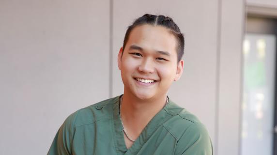 A smiling nursing student in green scrubs leans against a railing inside a modern building.