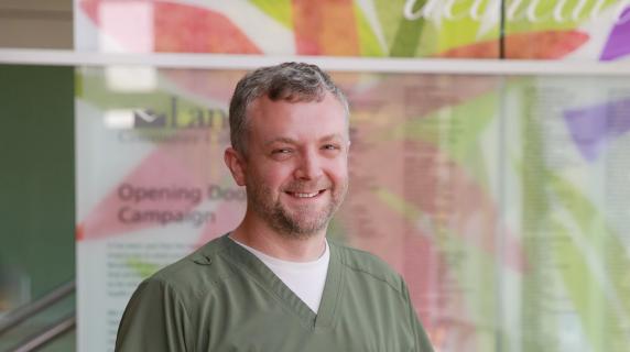 Stephen, a student in front of a mural wearing green scrubs over a white long  sleeve shirt and one hand in their pocket
