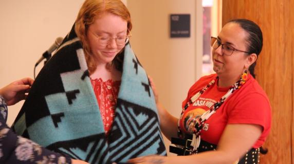 a native american young woman places a traditional blanket on another woman at a commencement ceremony in the Longhouse