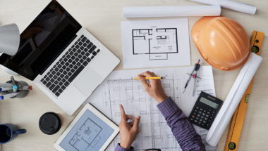 an engineer at a desk with a laptop, hard hat, and blueprints