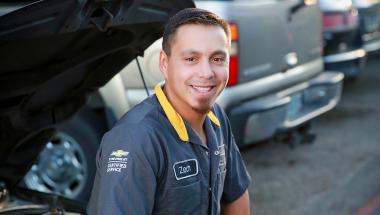 a scholarship winner, an automotive student posing next to an open car hood in his mechanic shirt