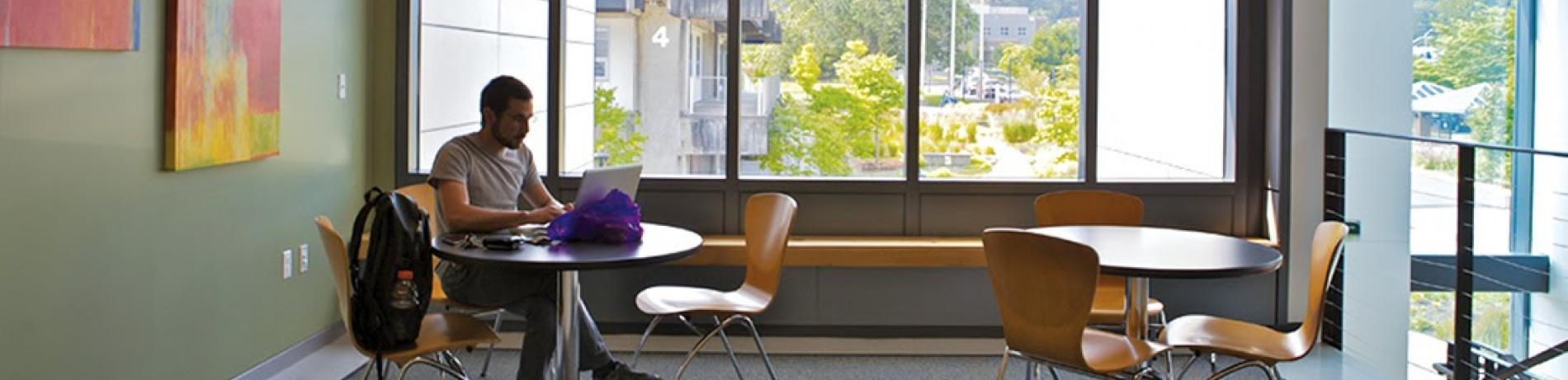 person sitting by a large window with a backpack working on a laptop