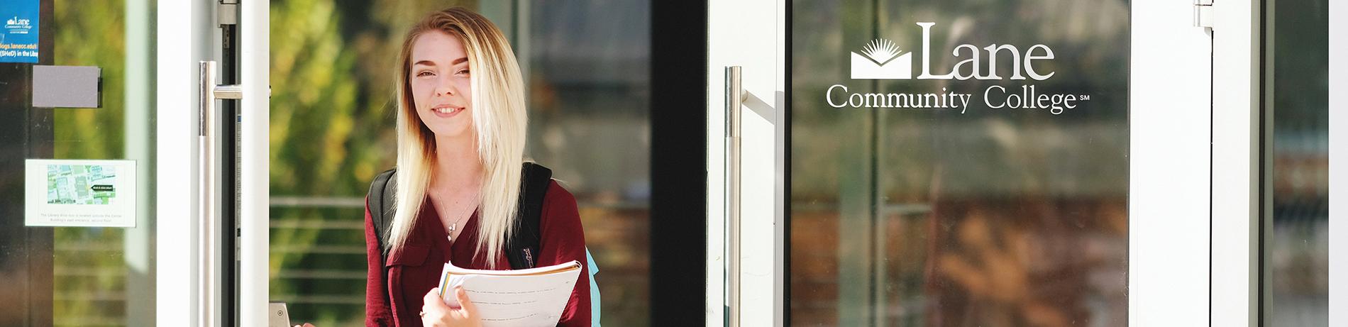 A student walks through a door smiling and holding notebook