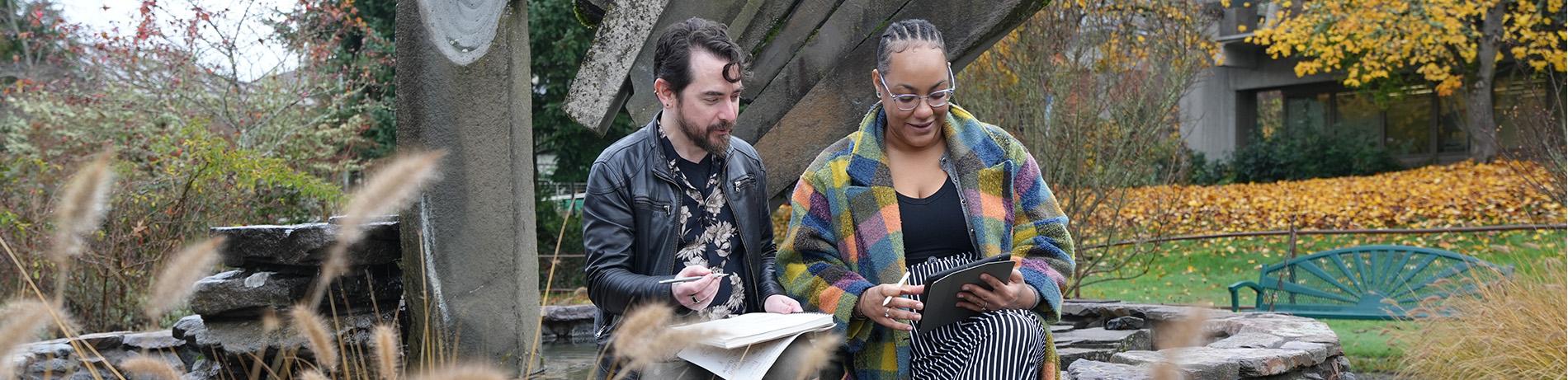 Two students, a white man in his 30s and a Black woman in her 20s, sit by the fountain on campus in autumn discuss school work