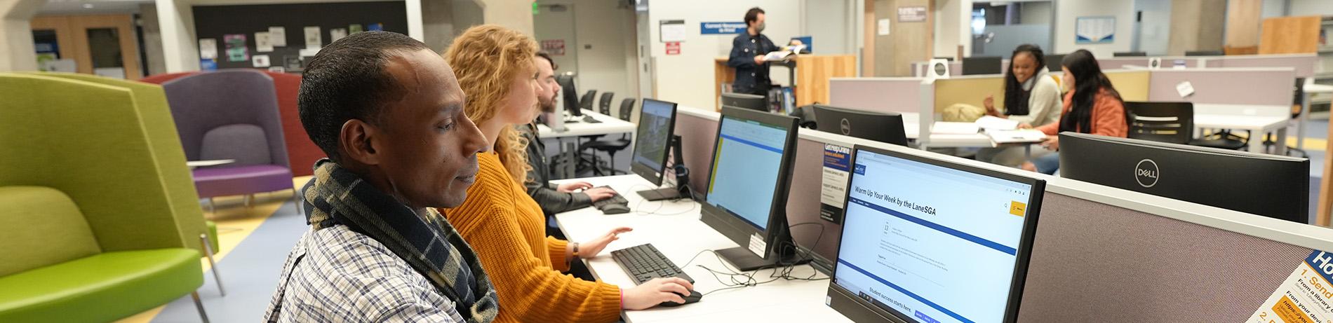 Diverse students working in the library. Two in the foreground work on computer, while three in the background research and study.