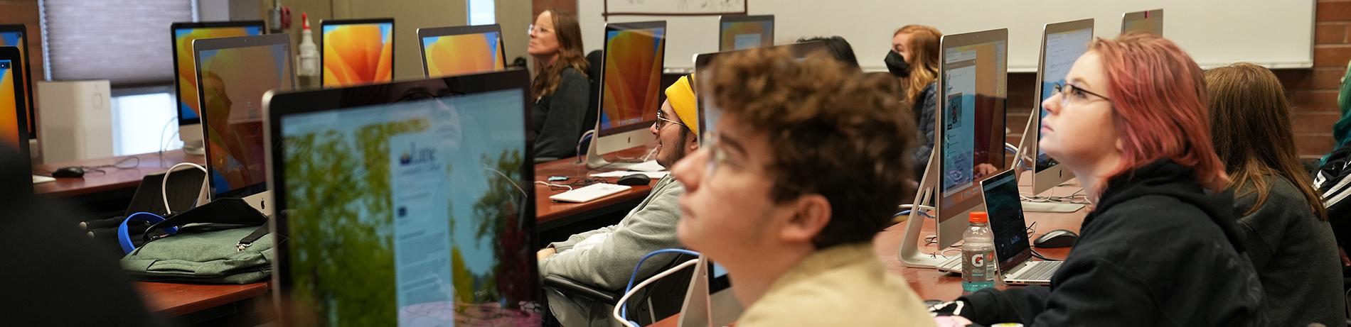 students in a computer lab listen attentively to a lecture by their instructor