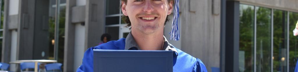 a graduating young man in blue cap and gown proudly shows off his LCC diploma