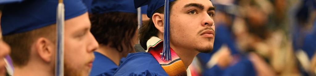 a graduating student, a Latino young man in a blue cap and gown and a serape stole, looks proudly in the distance during commencement