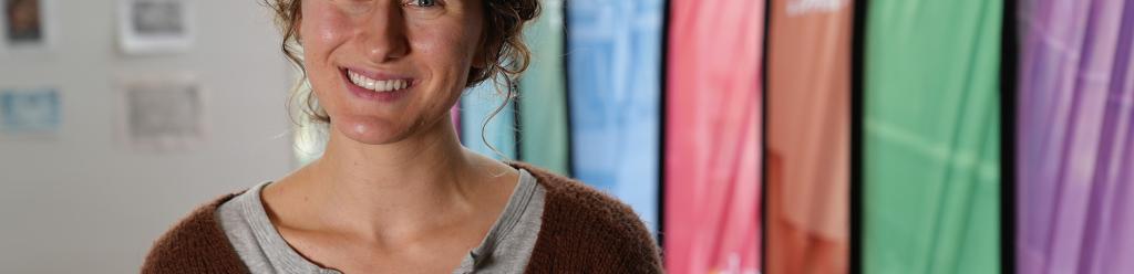 A student, a young woman with a gentle smile, wavy hair, and a brown cardigan, in the admissions building by colorful flags
