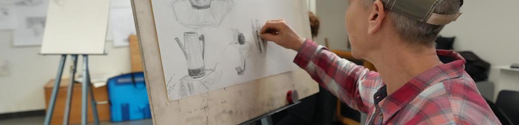 an art student, an older man with a flannel shirt and baseball cap, sketches a still life in a studio
