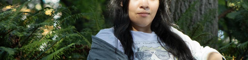 a young Latina woman on a bench with a cool shirt and jean jacket looks proudly at the camera with ferns and pines behind her