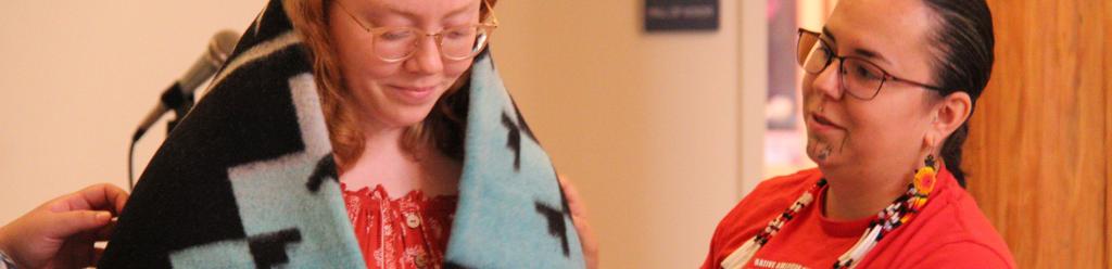 a Native American student with traditional face tattoos places a wool blanket on another Native American student in a ceremony in the Longhouse