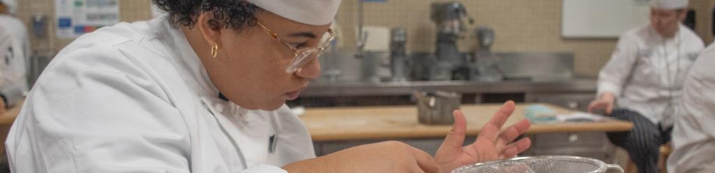 a culinary student, a Black woman in chef's clothes, concentrates as she sprinkles powder sugar in a kitchen